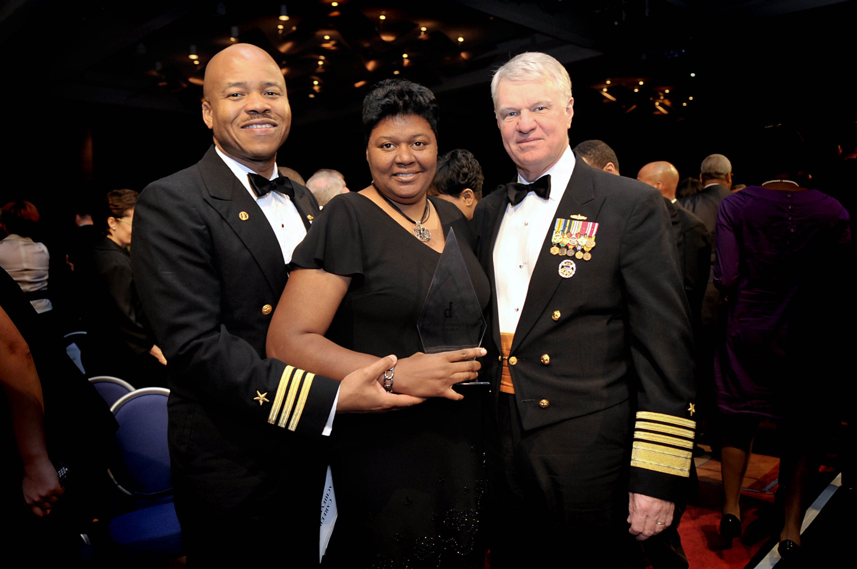 Zwei Männer in schwarzen Blazern und weißen Hemden stehen neben einer Frau in einem schwarzen Kleid, die eine Auszeichnung hält, auf einem roten Teppich, während andere sitzen und stehen und von beleuchteten Decken umgeben sind.