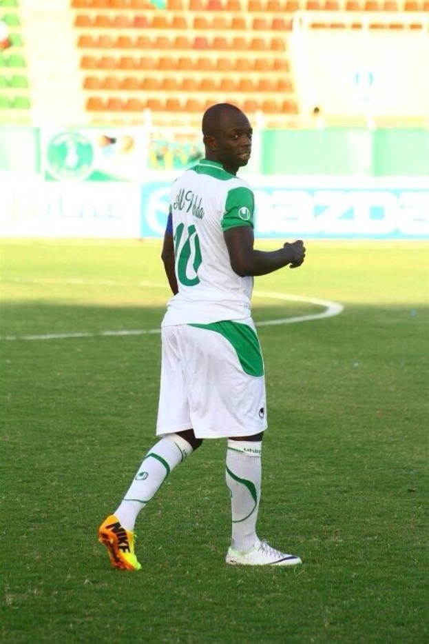 Ein Fußballer in grüner und weißer Uniform feiert auf einem Feld mit Werbetafeln und Stühlen während eines Spiels, wahrscheinlich nach einem Tor in der Afrikanischen Nationenmeisterschaft.