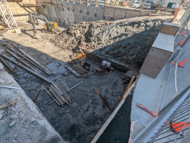 Baustelle mit einem großen Haufen Erde, umgeben von Kränen, Holzstangen und anderem Equipment, begrenzt von einem Maschendrahtzaun auf der rechten Seite, mit Gebäuden, Fahrzeugen, Pfählen, Bäumen und Himmel im Hintergrund.