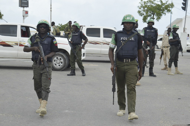Eine Gruppe von Polizisten in Helmen und mit Waffen auf der Straße gehend mit Fahrzeugen, Verkehrszeichen, Bäumen, Gebäuden und einem bewölkten Himmel im Hintergrund.