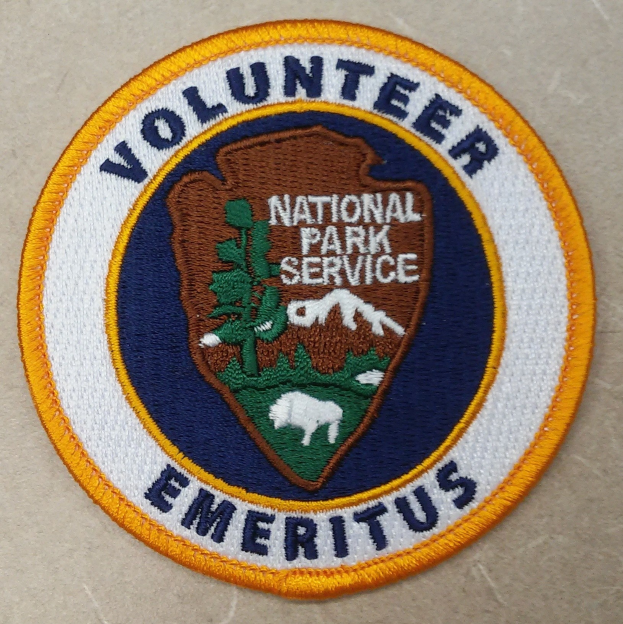 Runder Patch mit "Volunteer National Park Service Emeritus" in fetter schwarzer Schrift auf weißem Hintergrund, einem blauen Rand und einem gelben Kreis um den Text.