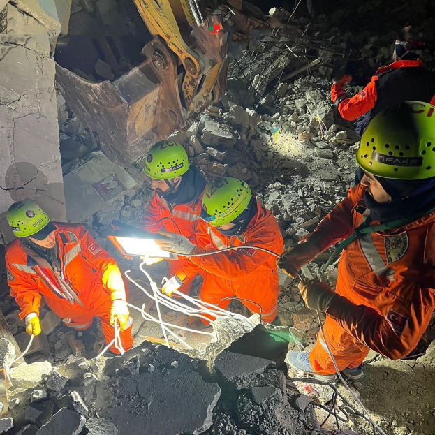 Gruppe von Menschen in orangefarbener Uniform und grüner Helm, die auf Schutt von einem eingestürzten Gebäude arbeiten, mit Werkzeugen und Materialien, mit einem Kran im Hintergrund.
