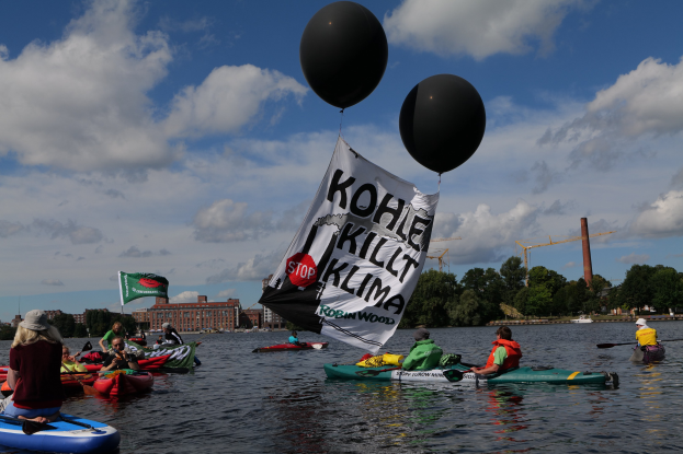 Gruppe von Menschen in Kajaks, die auf dem Wasser paddeln, mit einem Banner "Kohle Kill Klima" an einem der Boote befestigt, Bäume, Gebäude, Kräne und einen klaren blauen Himmel im Hintergrund.